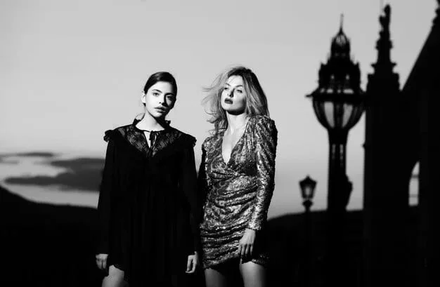 A black-and-white photograph of a Goth couple in 1980s London, featuring dramatic dark makeup, elaborate Victorian-inspired clothing like lace and velvet, and distinctive hairstyles, posed against a shadowy, atmospheric urban backdrop.