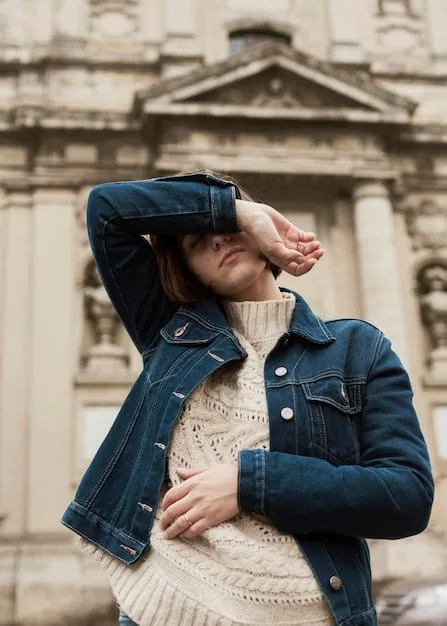 A person with a distinct, artistic sense of style, wearing an oversized, deconstructed denim jacket layered over a patterned dress, standing confidently in a brightly lit art gallery with abstract paintings in the background.