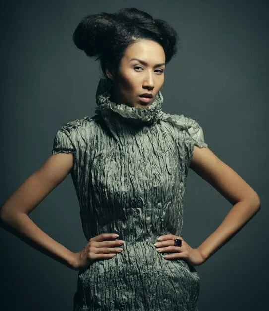 A fashion model is posing in a studio, wearing a dramatic outfit made from repurposed plastic bags and bottles. The outfit features intricate details and a futuristic silhouette, showcasing the potential of upcycled materials in high fashion.