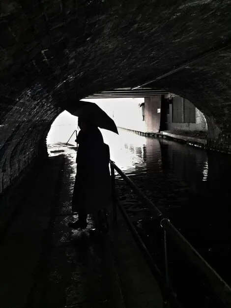 A black and white photo of an urban explorer standing in a flooded subway tunnel in New York City. They are wearing waders and holding a flashlight, illuminating the arched brick ceiling of the tunnel. The water reflects the tunnel's architecture, creating an eerie and symmetrical composition.