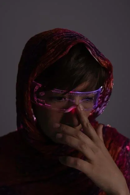 A gritty, close-up photograph of a person wearing a handmade cyberpunk mask with integrated LED lights and exposed wiring. The background is blurred, suggesting a crowded urban environment. Emphasis on the textures of the mask materials, such as plastic, metal, and electrical components.