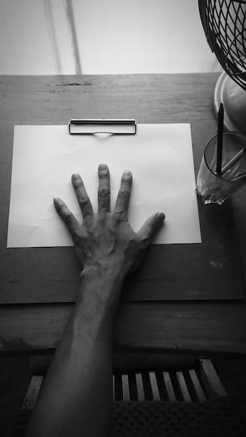 A black and white photograph of a weathered hand holding a pen, writing in a notebook. The hand is calloused, suggesting hard work.