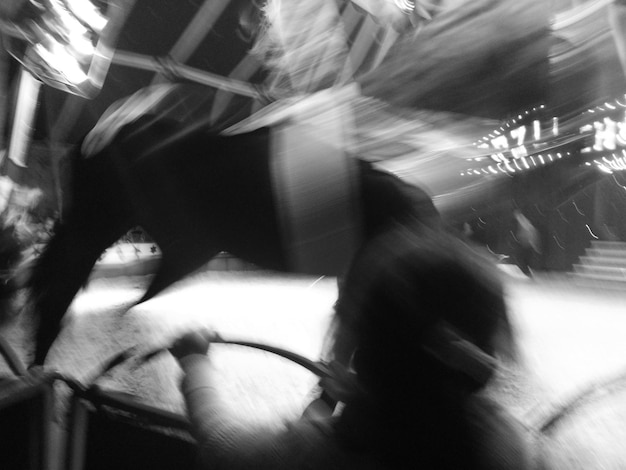 A black and white photograph of a DJ intensely mixing music at a dark underground club, with blurred figures dancing in the foreground and industrial-looking structures visible in the background.