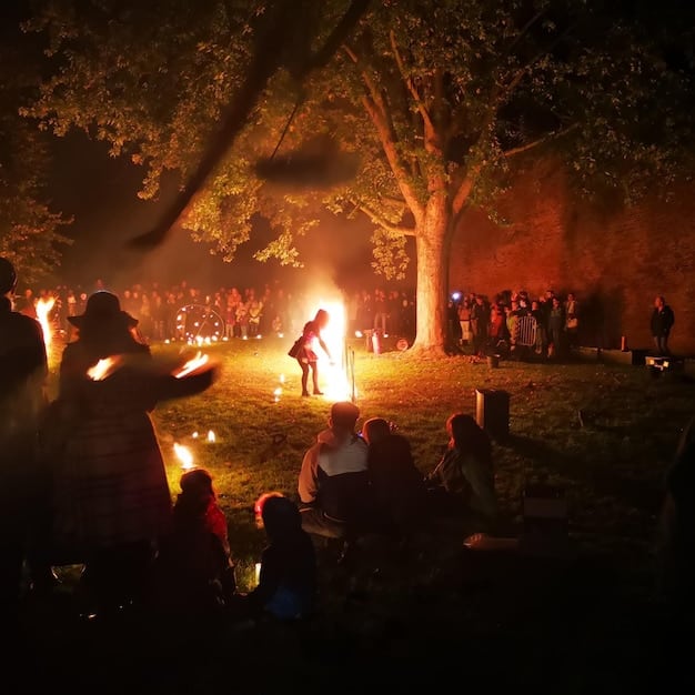 A group of people participating in a drum circle around a bonfire at night. The firelight illuminates their faces, and the rhythmic drumming creates a sense of community and spiritual connection.
