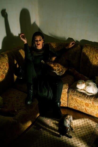 A young Goth individual sitting in a dimly lit room, illuminated by the glow of a laptop. They are designing digital art with Goth-inspired symbols and themes on the screen. The room is decorated with posters of Goth bands and dark, gothic elements.