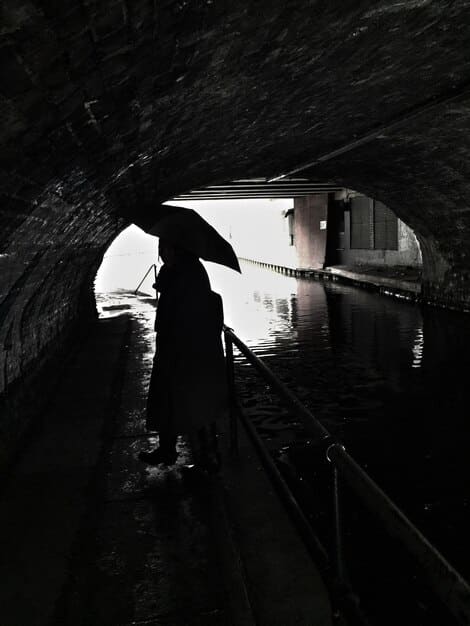 A black and white photo of an urban explorer standing in a flooded subway tunnel in New York City. They are wearing waders and holding a flashlight, illuminating the arched brick ceiling of the tunnel. The water reflects the tunnel's architecture, creating an eerie and symmetrical composition.