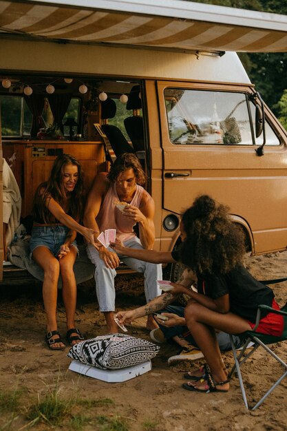 A group of diverse van dwellers gathered around a campfire in a remote campground, sharing stories and laughter. Several customized vans are parked in the background.
