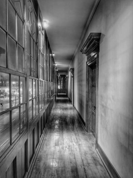 A black and white photo of an empty, decaying library inside an old, abandoned university building somewhere in the US. Books are scattered on the floor and shelves, and dust particles float in the air illuminated by sunlight piercing through cracks in the boarded-up windows. The photo evokes a sense of lost knowledge and the passage of time.