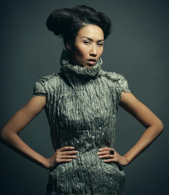 A fashion model is posing in a studio, wearing a dramatic outfit made from repurposed plastic bags and bottles. The outfit features intricate details and a futuristic silhouette, showcasing the potential of upcycled materials in high fashion.