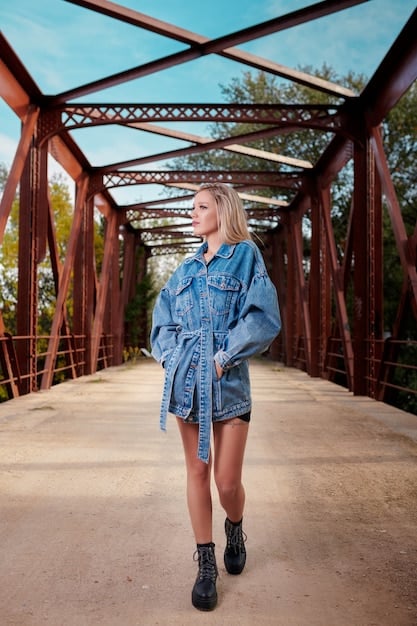 Full-length shot of a person wearing a deconstructed denim jacket with multiple pockets, raw edges, and contrasting denim washes. They are standing in an urban alleyway with graffiti art in the background, radiating confidence and effortless cool.