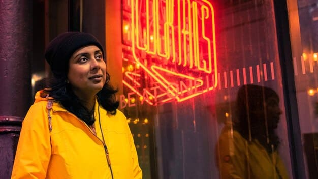 A street-style photograph featuring a person wearing a bright red vinyl jacket over a neon yellow graphic tee, paired with plaid pants in shades of blue and green. They are accessorized with chunky silver jewelry and platform boots. The backdrop is a graffiti-covered wall, emphasizing the rebellious and eclectic nature of underground fashion.
