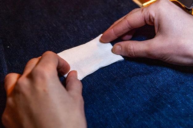 A close-up shot of hands sewing a patch onto a denim jacket. The jacket is heavily customized with various patches, studs, and hand-painted designs, showcasing the DIY nature of underground fashion. The background is a cluttered workbench with sewing tools and fabric scraps.