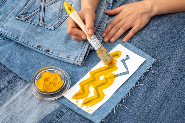 A close-up of a designer's hands adding intricate patchwork details to an upcycled denim jacket, showcasing the meticulous craftsmanship involved in DIY fashion. Threads and fabric scraps are visible on a workbench.