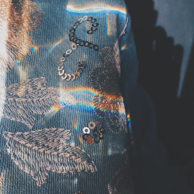 A close-up of a designer's hand meticulously sewing intricate details onto a unique, avant-garde garment. Fabric swatches and patterns are visible in the background.